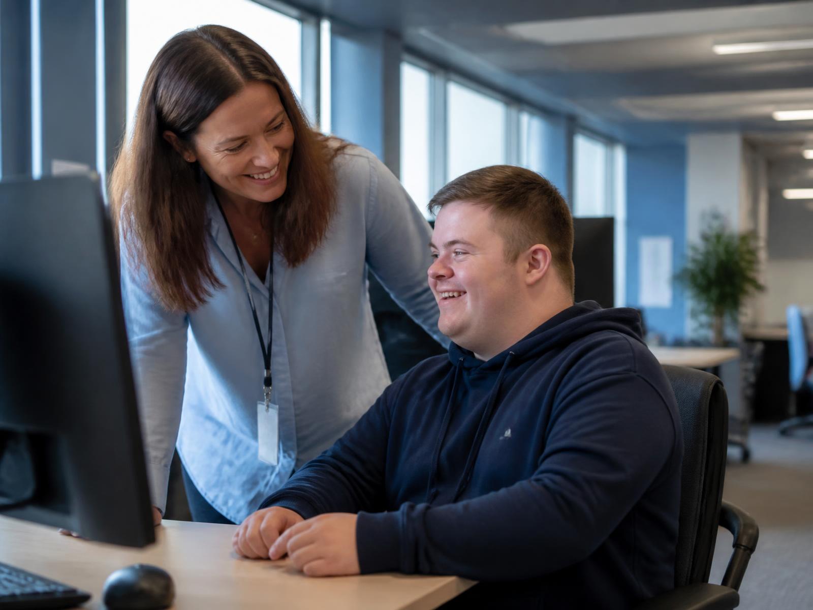 An employment specialist smiling alongside a young man with Down syndrome at his workplace desk